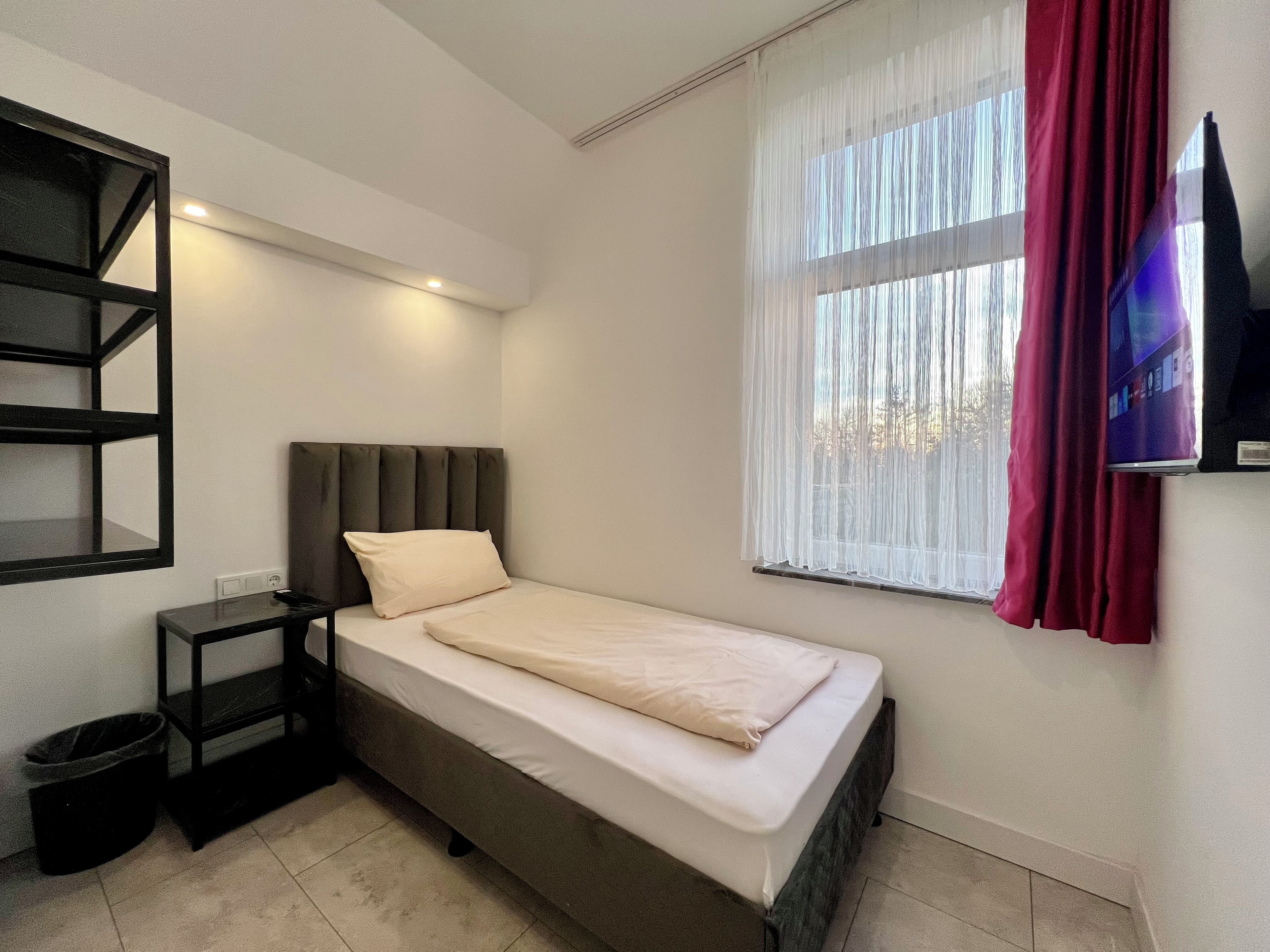 Una cama individual con cabecera, una mesita de noche, un televisor montado en la pared y una ventana con cortinas.
