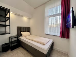 A single bed with a headboard, a bedside table, a TV mounted on the wall, and a window with curtains. - Durmaz Hotel (Hannover)