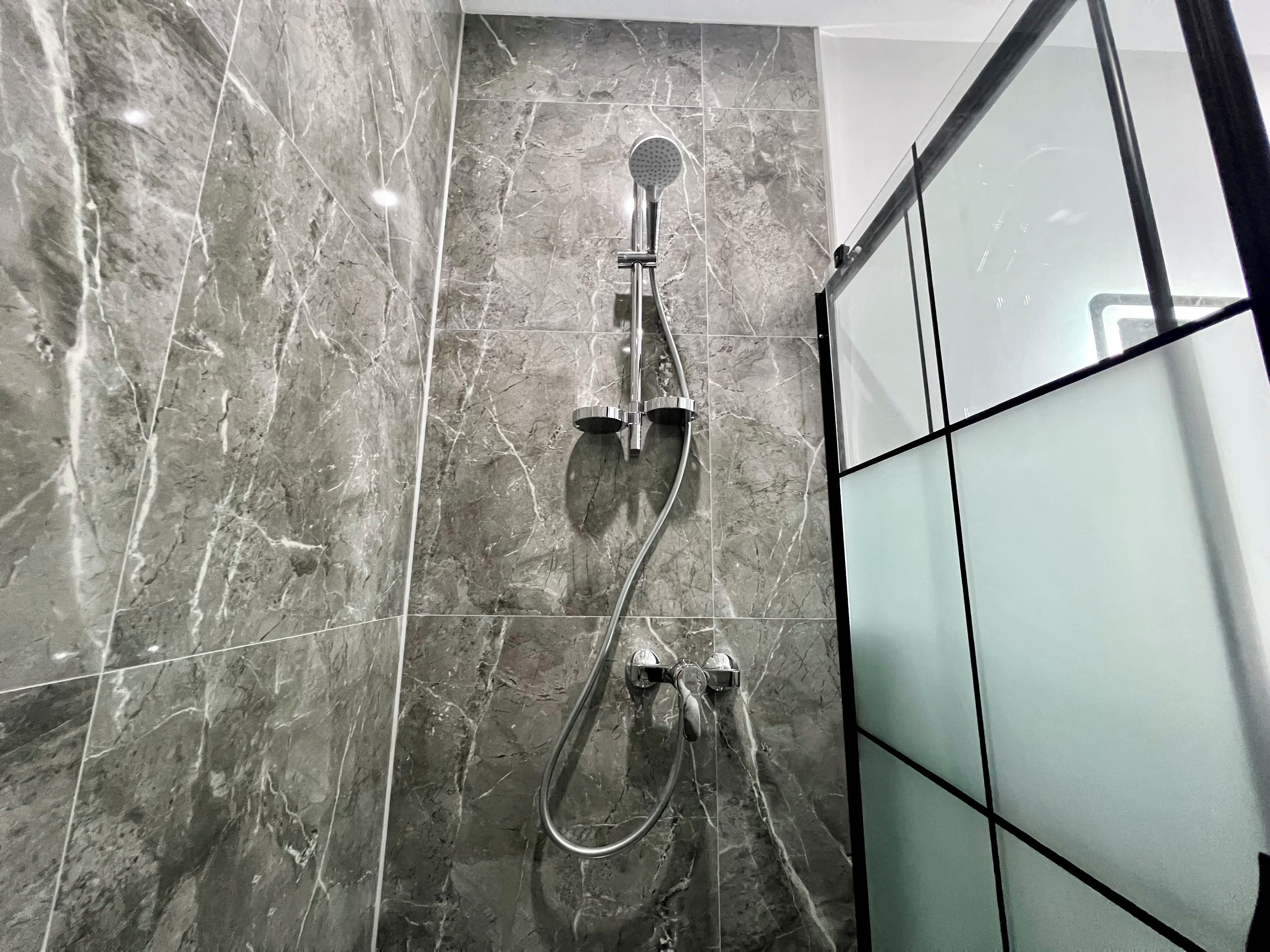 A shower with a handheld showerhead and a wall-mounted faucet, set against a marble wall.
