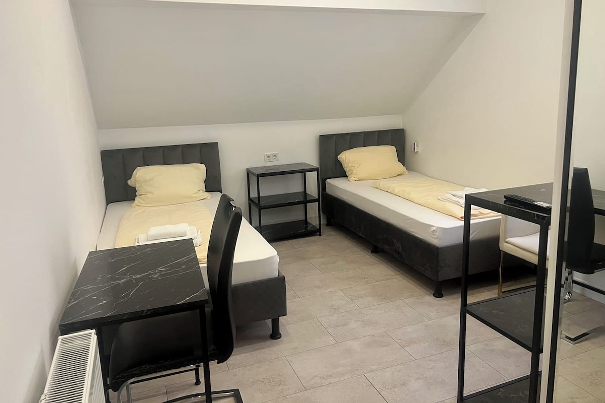 A compact hotel room with two single beds, a desk with a chair, a small table, and a radiator.