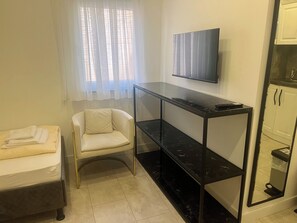 Een kleine, goed verlichte kamer met een bed, een stoel, een aan de muur bevestigde televisie en een console tafel.