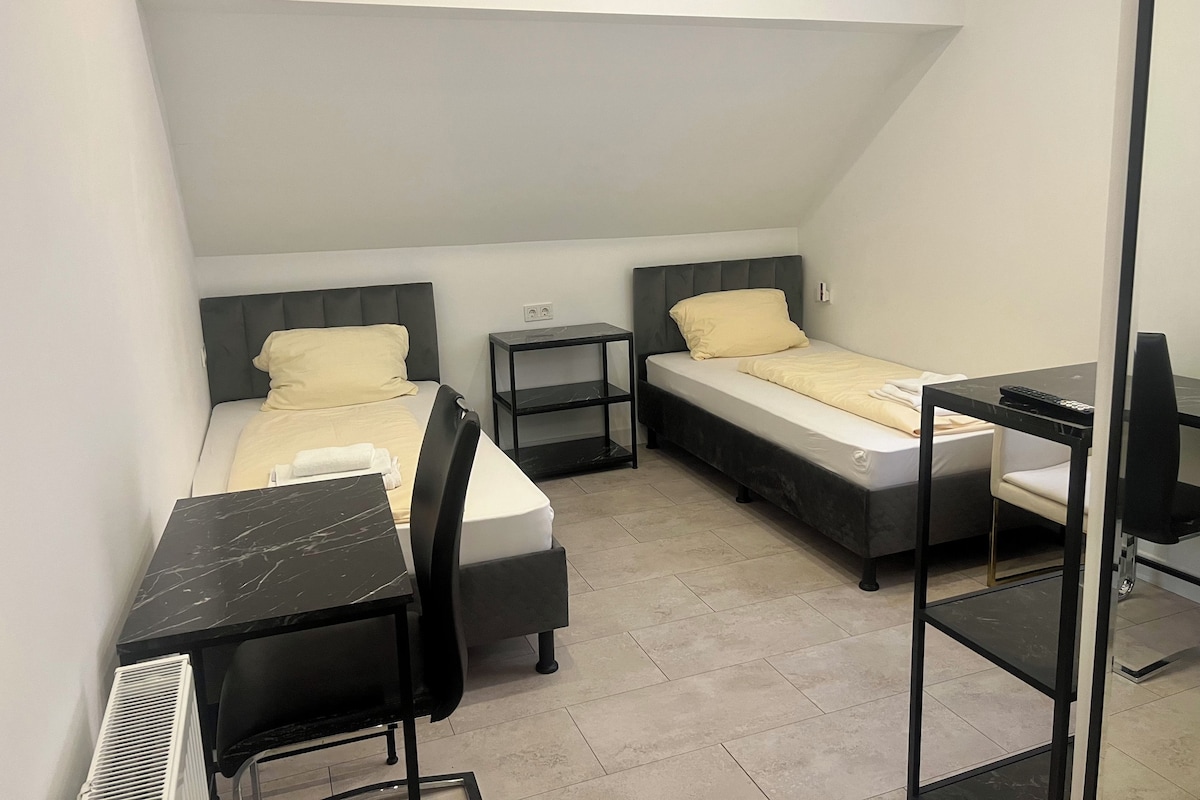 Et kompakt hotelværelse med to senge, et skrivebord med stol, et lille bord og en radiator.