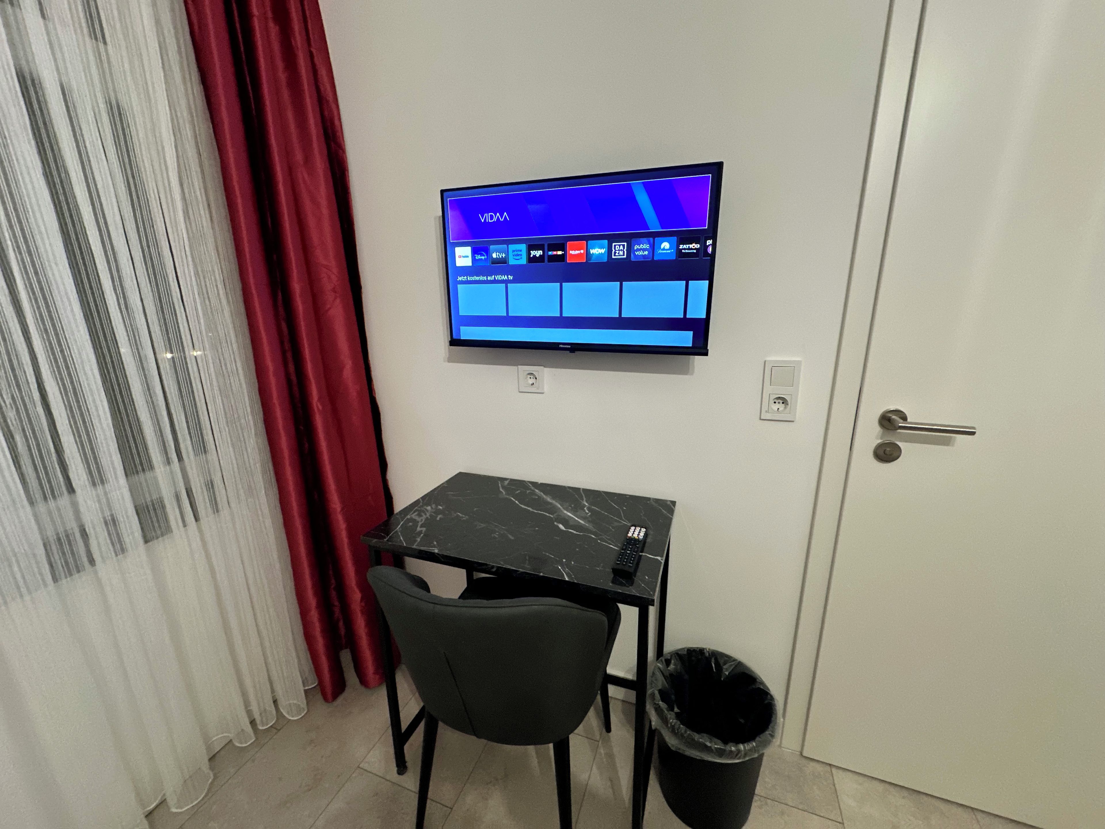 A room with a TV mounted on the wall, a small table with a chair, a door, and a trash bin.
