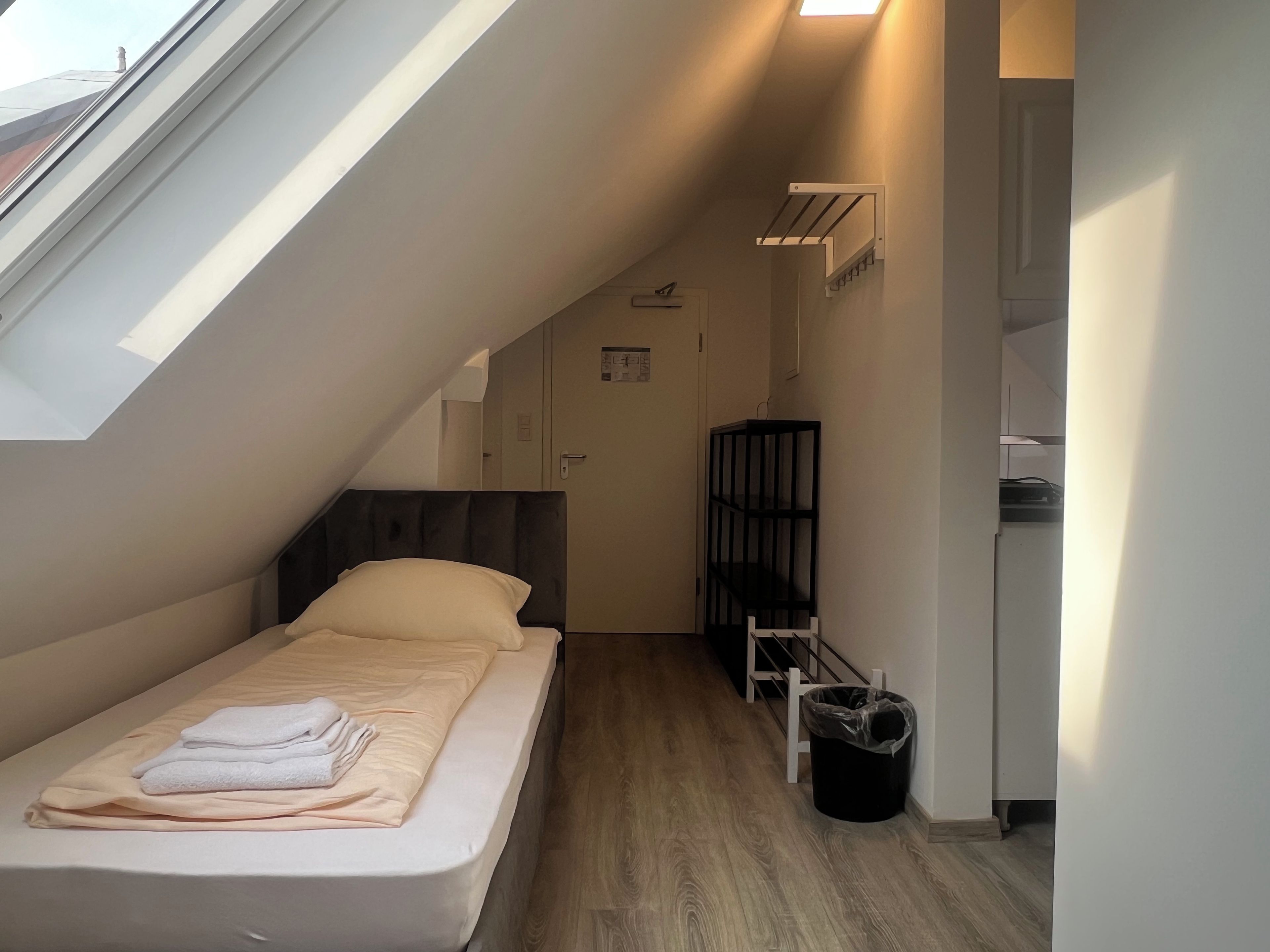 A compact room with a bed, a small trash bin, and a black shelving unit.