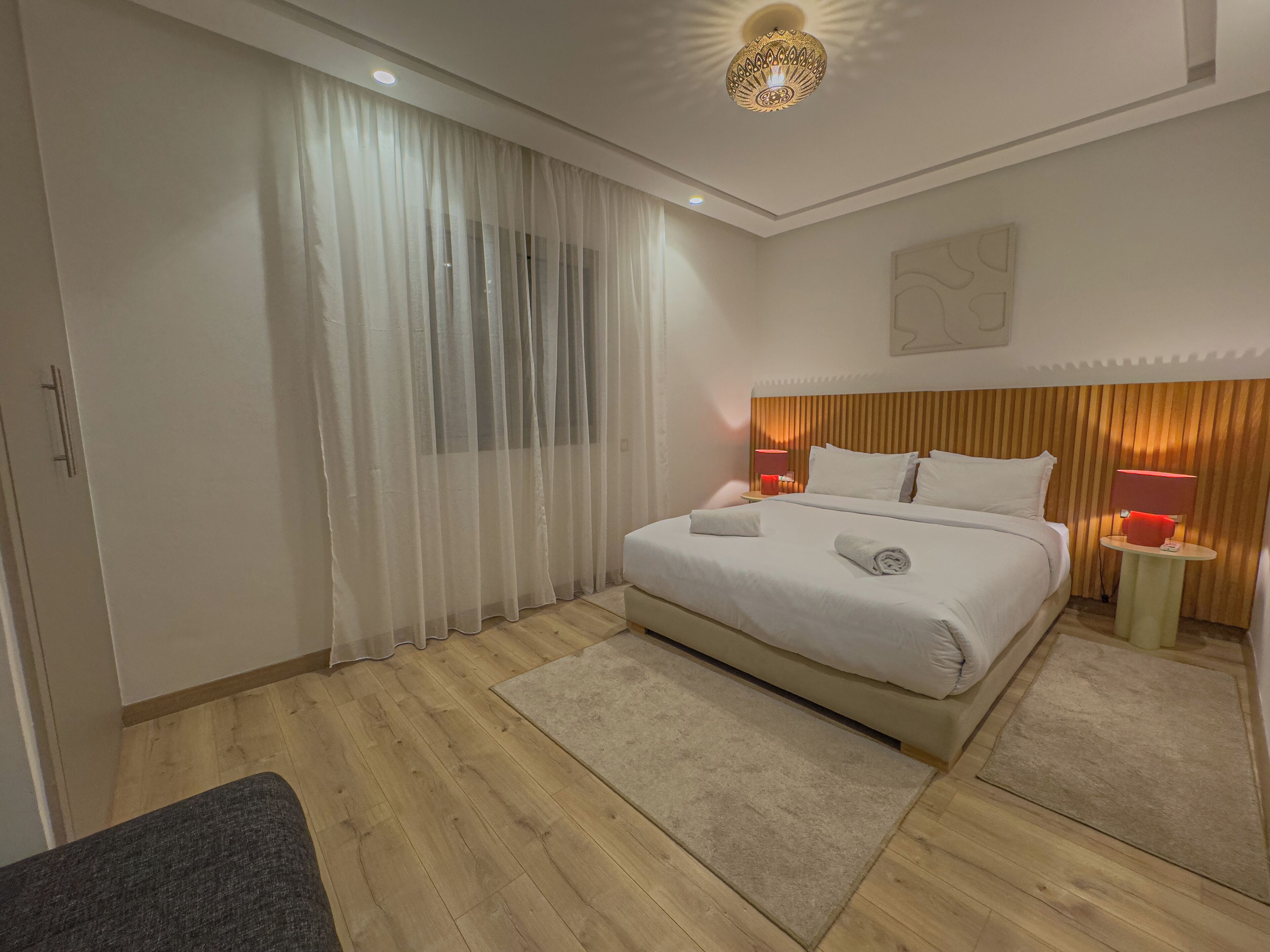 A modern hotel room with a large bed, a wooden headboard, a bedside table with a lamp, and sheer curtains.