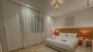 Une chambre d’hôtel moderne avec un grand lit, une tête de lit en bois, une table de chevet avec une lampe et des rideaux en voile.