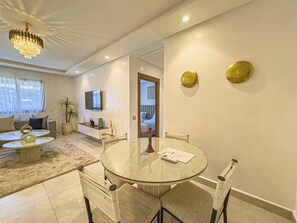 Una sala de estar moderna con una mesa redonda de vidrio, sillas acolchadas, un sofá, un televisor y elementos decorativos en la pared.