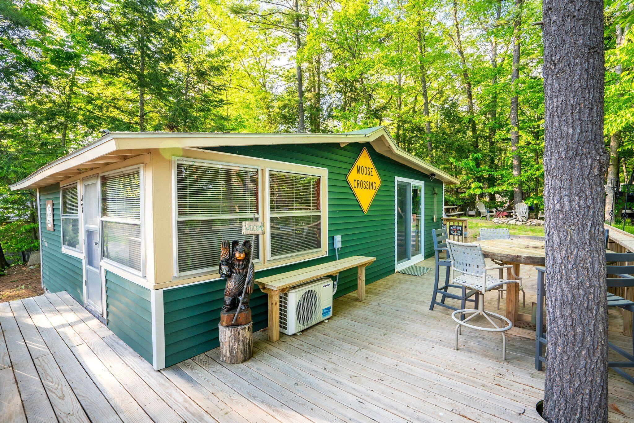 Una cabaña verde con un letrero que dice 'Moose Crossing', una terraza de madera y mobiliario exterior.