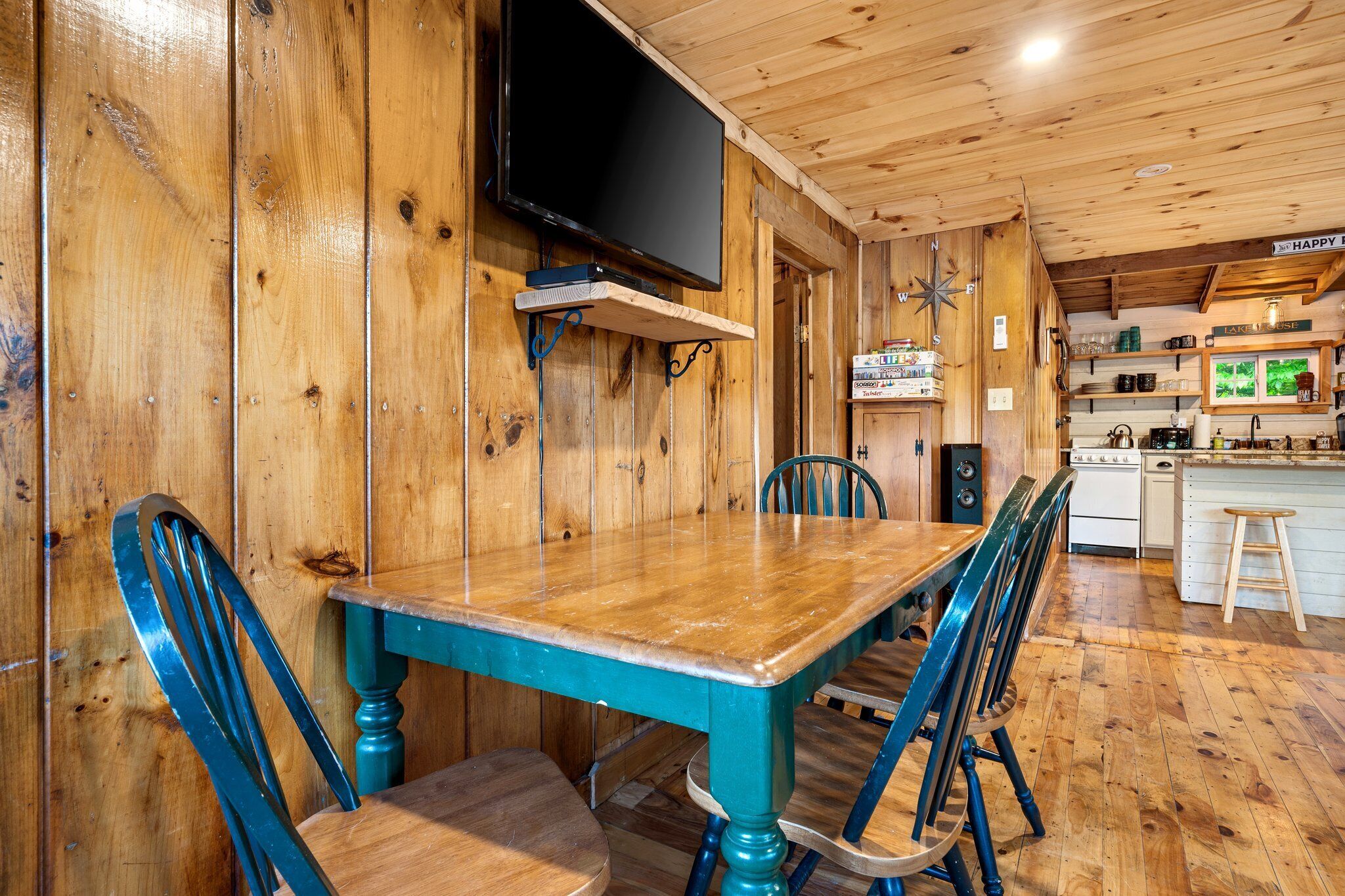 Una mesa de comedor de madera con sillas, un televisor montado en la pared y una cocina con alacenas y electrodomésticos.
