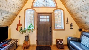 A cozy attic living room with a wooden sloped ceiling, a dark wooden door, and two windows with autumn foliage views. - Canyon A-Frame Cabin | Fire Pit, Sauna, Hiking (Fort Payne)