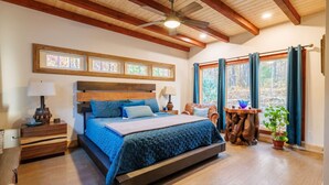 A bedroom with a large bed, wooden headboard, and a ceiling fan. There are windows with curtains, a nightstand, and a small table with a plant. - Canyon A-Frame Cabin | Fire Pit, Sauna, Hiking (Fort Payne)