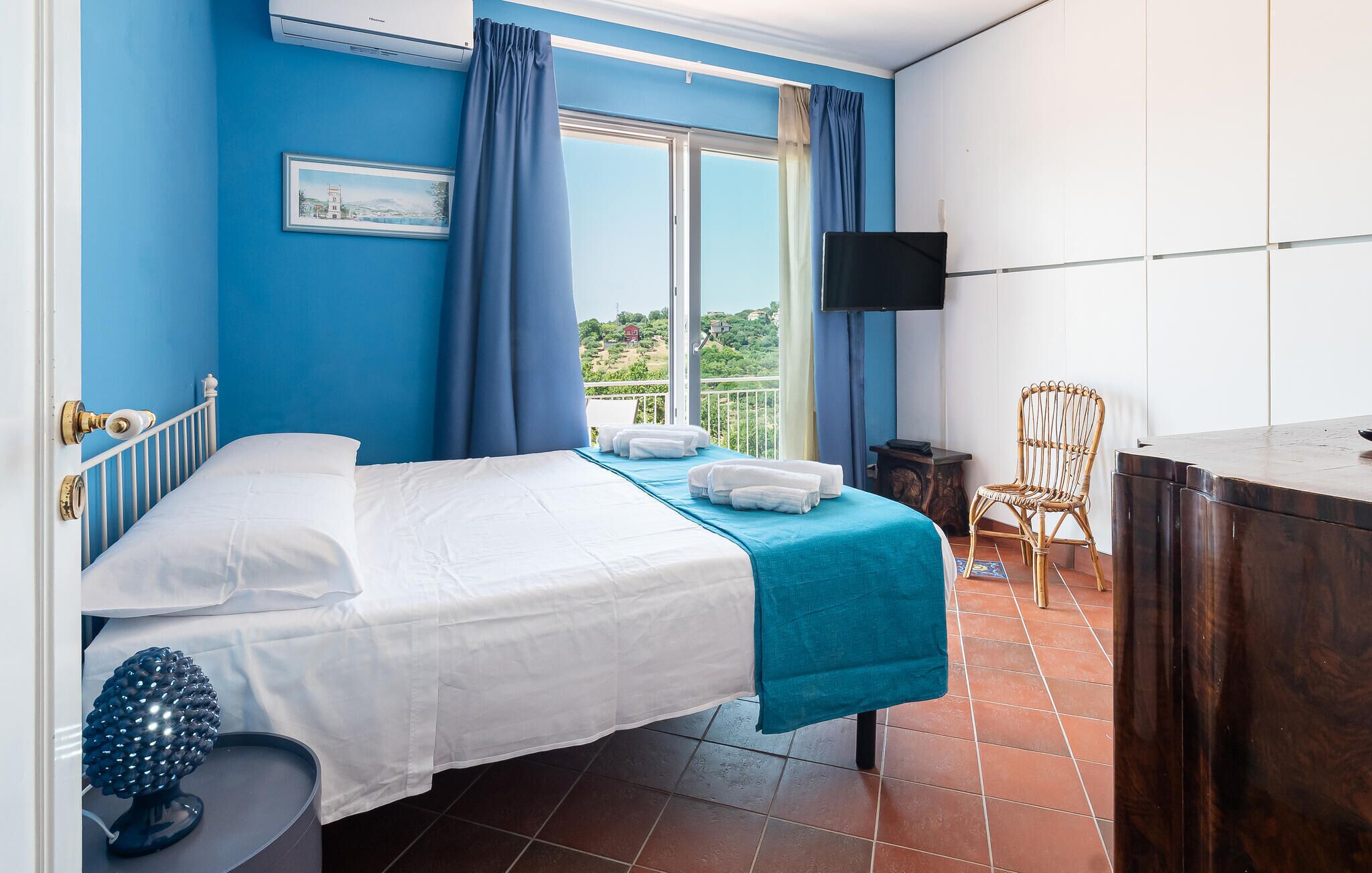 A hotel room with a bed, a TV, a chair, and a balcony with a view.