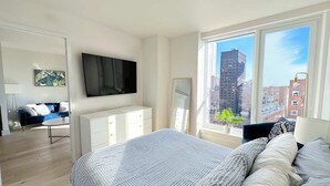 A modern bedroom with a large bed, a dresser, a TV, and a view of the city through large windows. - Luxury 1BR - Balcony- Elevator - Gym - Lounge -Rooftop -In Unit Laundry - Manhattan (New York)