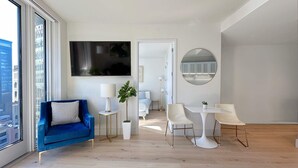 A modern living room with a blue sofa, a dining table with white chairs, a TV mounted on the wall, and a large window with a view of buildings. - Luxury 1BR - Balcony- Elevator - Gym - Lounge -Rooftop -In Unit Laundry - Manhattan (New York)