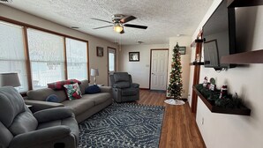 Un salon avec un canapé gris, un fauteuil gris, un plancher en bois, un ventilateur de plafond et un sapin de Noël.