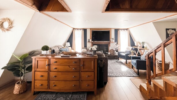 A loft-style living space with a wooden staircase, a fireplace, a sofa, and a dining area. - Cozy Wister Ridge A-Frame Lake Cabin • Sleeps 6 • Mins to Wister Lake! (Wister)