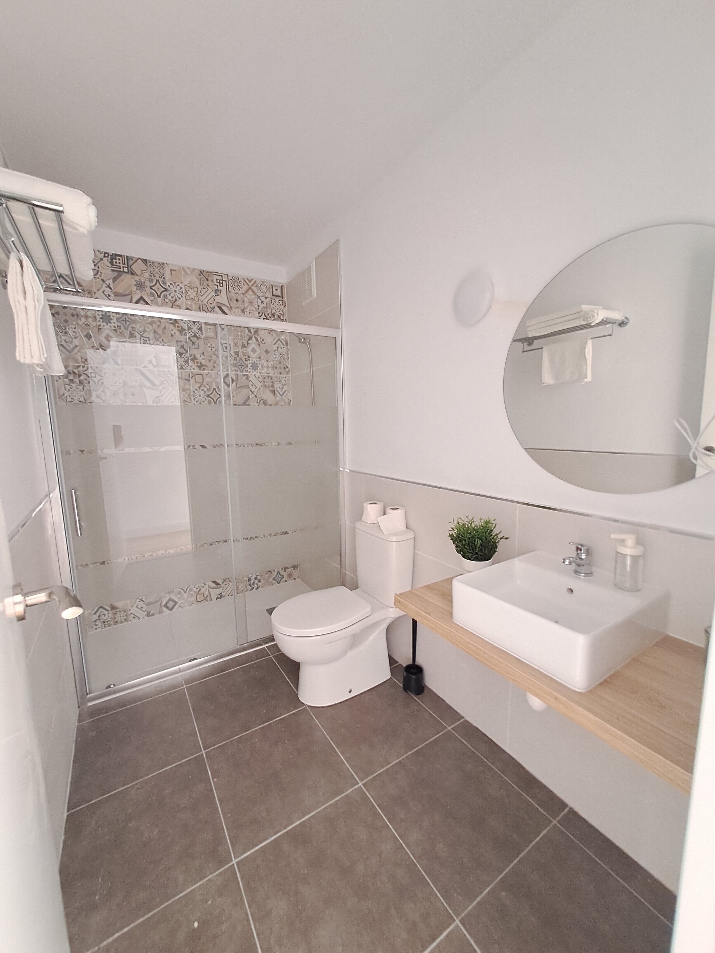 Un bany modern amb plat de dutxa envoltat de vidre, un vàter, un lavabo amb mirall i una planta en test.