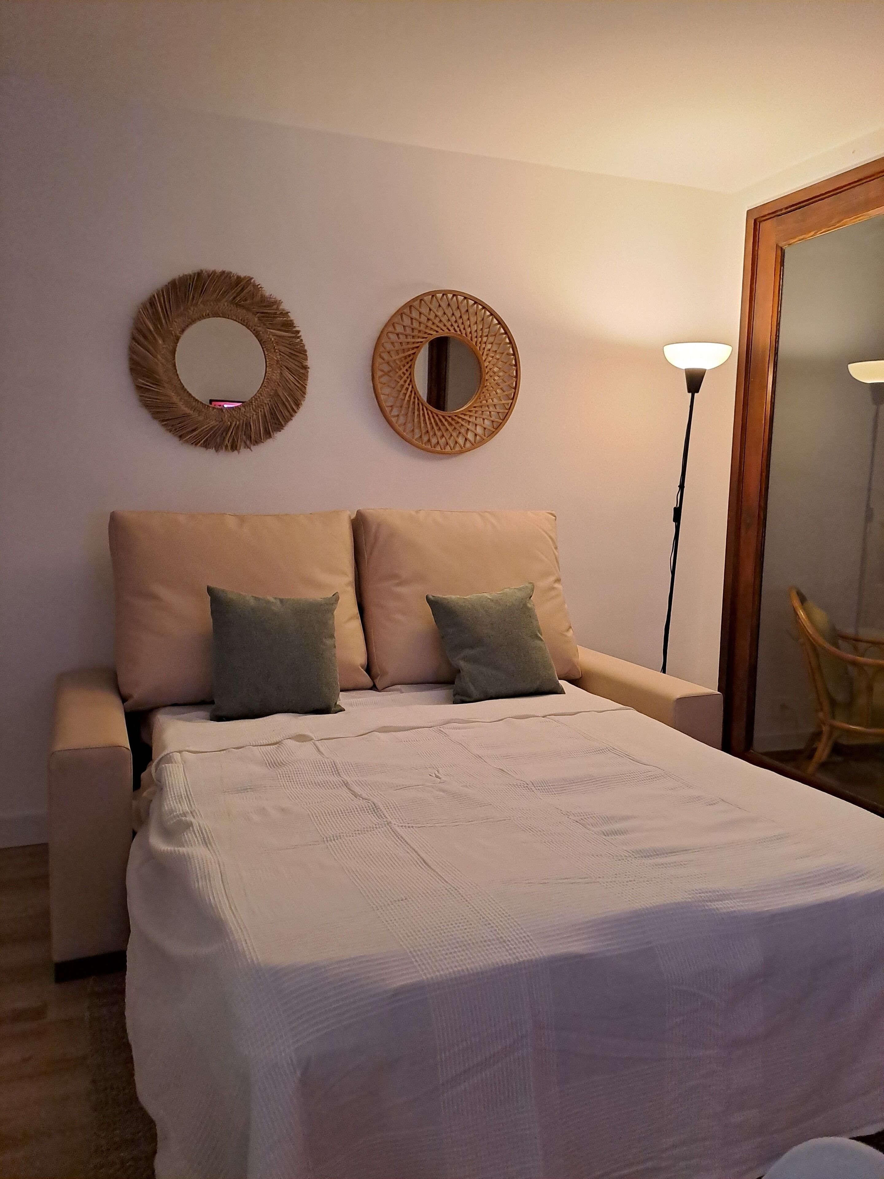A bedroom with a bed, two pillows, a bedside table, a floor lamp, and two wall mirrors.