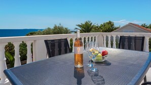 Une table avec une bouteille de vin, des verres et un saladier de fruits, sur un balcon donnant sur la mer.