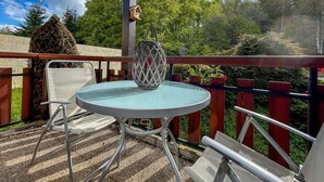 Une table ronde avec un plateau en verre, deux chaises blanches, une lanterne suspendue et une nichoir en bois sur un balcon offrant une vue sur les arbres et un ciel dégagé.
