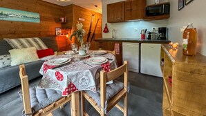 Une pièce chaleureuse avec une table à manger en bois, préparée pour quatre personnes, un canapé gris agrémenté d’un coussin rouge, des meubles en bois, un four à micro-ondes et un petit coin cuisine.