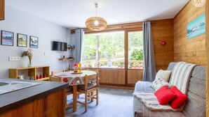 Une pièce chaleureuse avec un décor intérieur en bois, une table à manger avec des chaises, un canapé agrémenté d’un coussin rouge et une grande fenêtre donnant sur un paysage verdoyant.