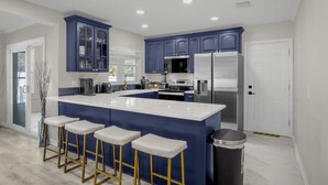 A modern kitchen with blue cabinets, a white island with bar stools, stainless steel appliances, and a trash can. - Luxury 6BR Villa with Heated Pool & Private Theater (Valrico)