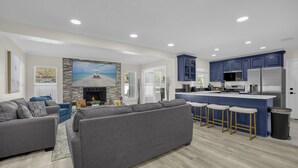 A modern living room with a gray sofa and a kitchen area featuring blue cabinets and a stainless steel refrigerator. - Luxury 6BR Villa with Heated Pool & Private Theater (Valrico)