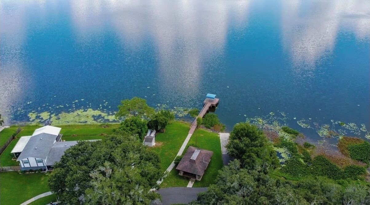 An aerial view of a residential area with a dock extending into a lake, surrounded by lush greenery and houses.