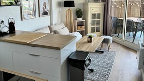 Una cocina moderna con encimera de madera, lavavajillas empotrado y cubo de basura negro. Hay una zona de comedor con mesa y sillas de madera, una pequeña planta en maceta y una alfombra con estampado blanco y negro. Un ventanal ofrece vistas al exterior.