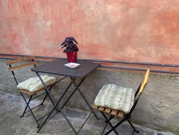 A small outdoor table with a red pot and a white bowl, two foldable chairs with patterned cushions, and a stone floor.