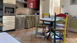 A kitchen with a stove, oven, and dishwasher, a dining table with colorful chairs, and a window with curtains. - The Pink House of Riso Amaro In the painted village Legro  of Orta San Giulio (Orta San Giulio)