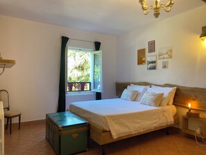 Une chambre à coucher avec un lit, une valise, une fenêtre avec des rideaux, un lustre et des œuvres d’art murales.