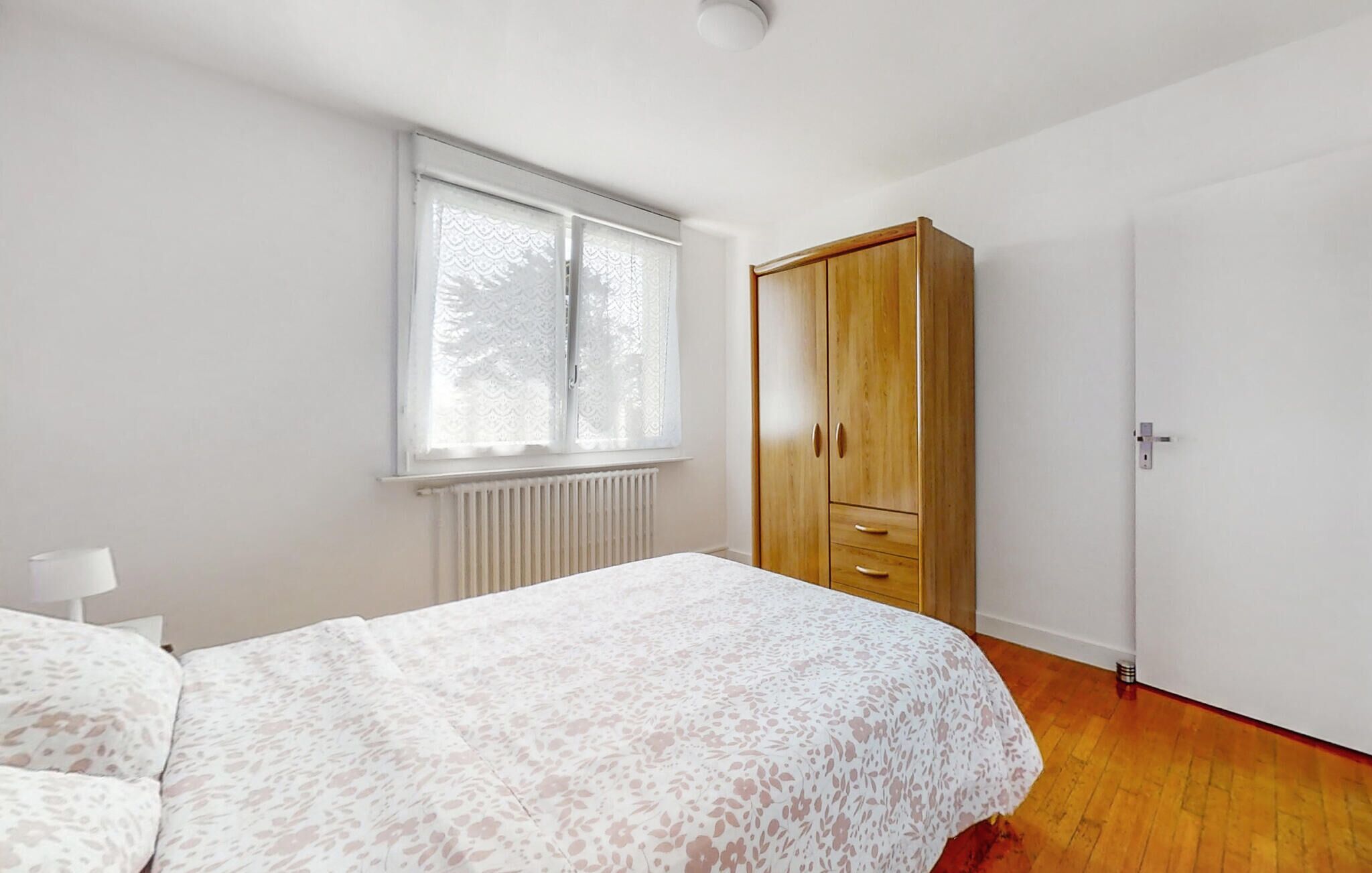 Ein Schlafzimmer mit Bett, Fenster mit Spitzenvorhängen, einem Holzschrank und einem Heizkörper.