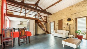 Ein großzügiger Wohnbereich mit hoher Decke, freiliegenden Holzbalken, einer Treppe, einem Essbereich mit rotem Tisch und Stühlen, einem weißen Sofa und einem Couchtisch.