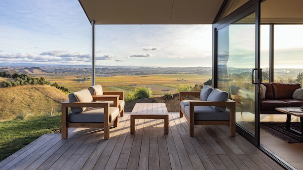 A modern outdoor deck with wooden furniture and glass walls offering a scenic view of rolling hills.