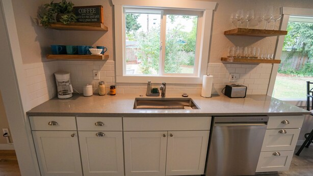 Una cocina con armarios blancos, lavavajillas de acero inoxidable, cafetera y estantes con tazas y vasos.