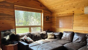 L’intérieur chaleureux d’un chalet avec un grand plafond en bois, un canapé en cuir agrémenté de coussins, un téléviseur posé sur un meuble en bois et une fenêtre équipée de stores.