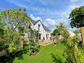 Una casa moderna de dos pisos con un césped bien cuidado, una terraza de madera y una variedad de plantas en macetas.