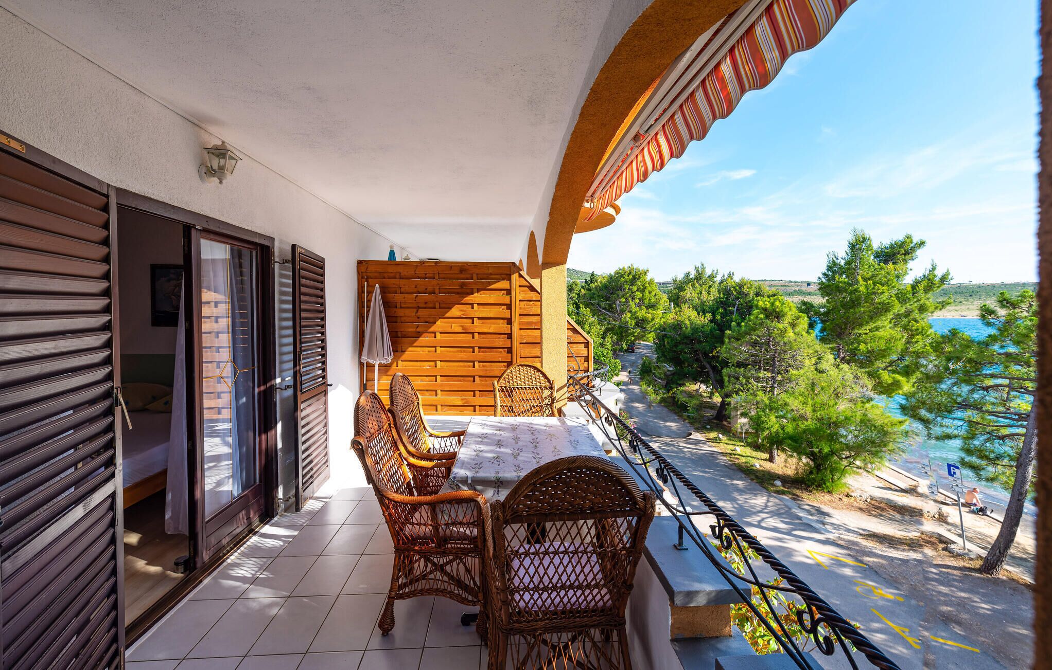 A balcony with wicker chairs and a table, overlooking a coastal area with trees and a beach.