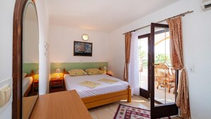 Une chambre à coucher avec un lit, des tables de chevet, une horloge, un tableau et une vue sur l’extérieur, visible à travers une porte ouverte.
