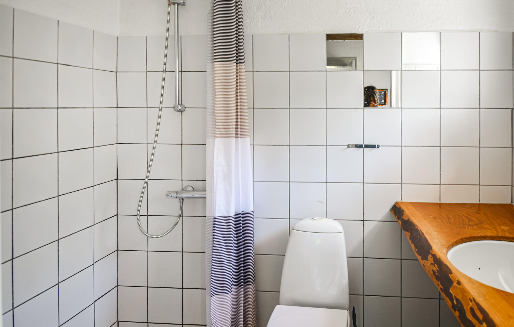 A bathroom with white tiled walls, a shower with a striped curtain, a toilet, and a wooden vanity with a sink.