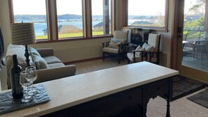 A cozy living room with a sofa, two armchairs, a coffee table, and a TV. Large windows offer a view of a lake and buildings. - Ridge View Suite Seascape Panorama with Covered Deck  (Campbell River)