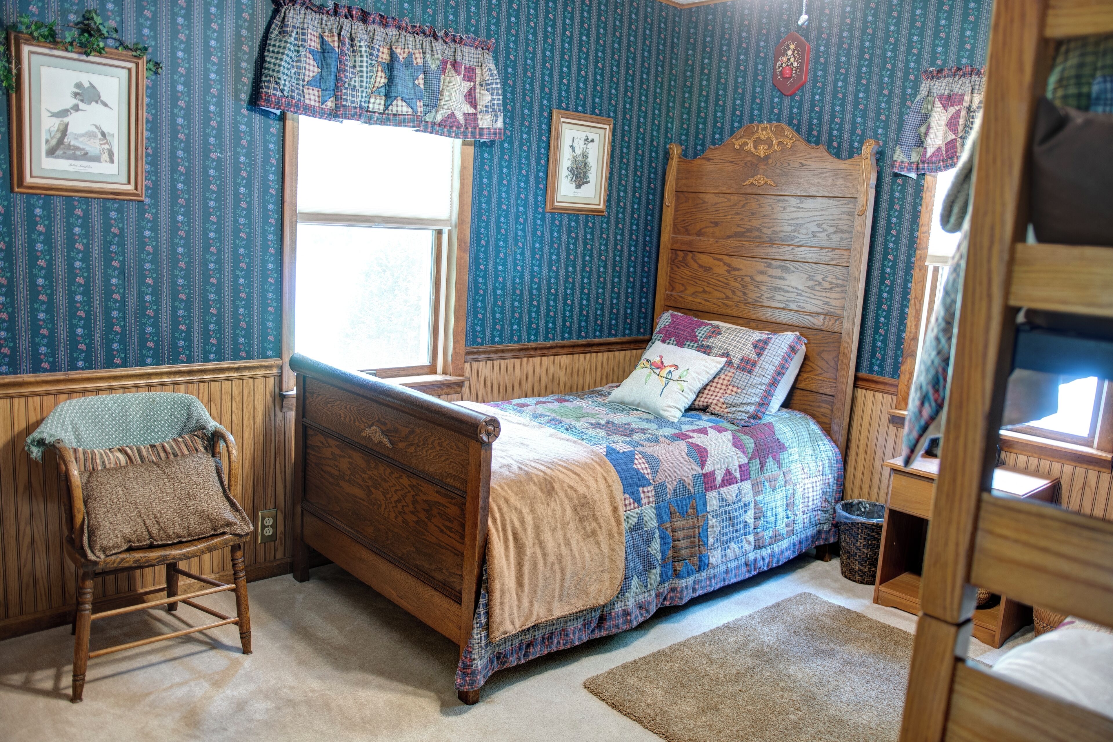 A bedroom with a wooden bed, a chair, a window, and patterned wallpaper.