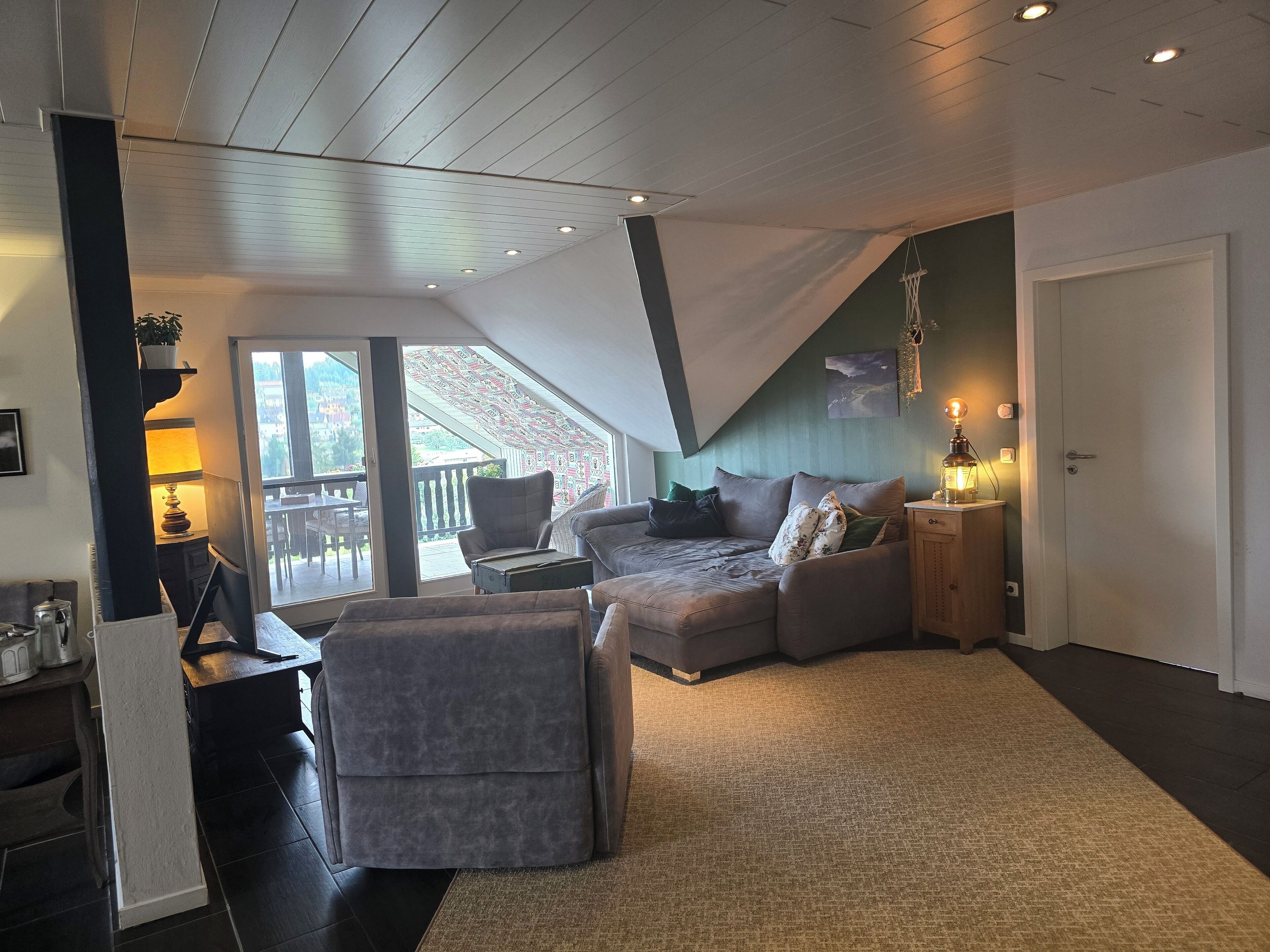Ein gemütliches Wohnzimmer mit einer Couch, Sesseln und einem kleinen Tisch mit Lampe. Ein Schiebeglastür führt zu einem Balkon.