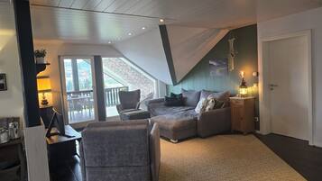 Ein gemütliches Wohnzimmer mit einer Couch, Sesseln und einem kleinen Tisch mit Lampe. Ein Schiebeglastür führt zu einem Balkon.