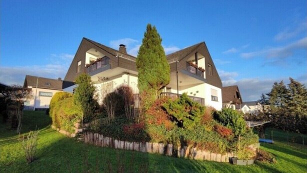 Uma casa de dois andares com telhado inclinado, varanda no andar superior e um jardim bem cuidado com arbustos diversos e uma árvore alta.