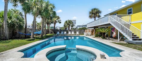 A swimming pool with a hot tub, surrounded by palm trees and a yellow building with a white staircase.