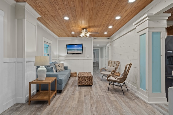 A modern living room with a wooden ceiling, a blue sofa, wicker chairs, and a television mounted on the wall. - Sunshine Oasis | Private Pool | Dog-Friendly | My Beach Getaways (Gulf Breeze)