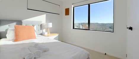 A hotel room with a bed, a nightstand, a lamp, a window with a view, and a door.